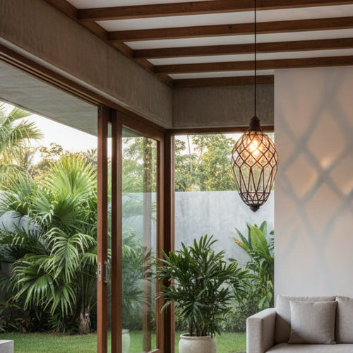A modern pendant light with a black brass frame and warm glowing bulb hangs in a minimalist living room with a light gray sofa, wooden ceiling beams, and large glass doors opening to a lush green outdoor patio.
